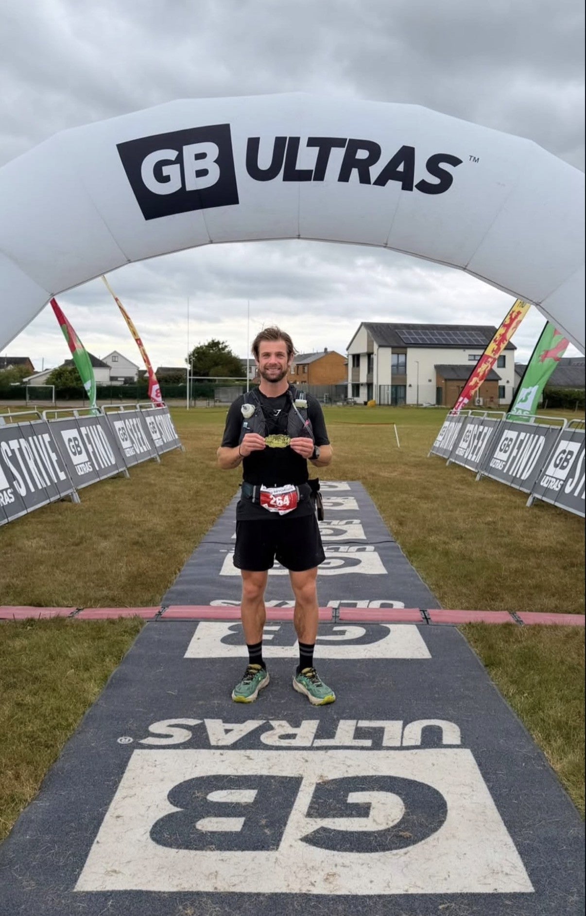 Contact Coffee Co founder Luke Dalton at the Beacons Way Ultra 100 finish line in Llangadog, standing under the GB Ultras arch in bib 264 holding his finisher buckle